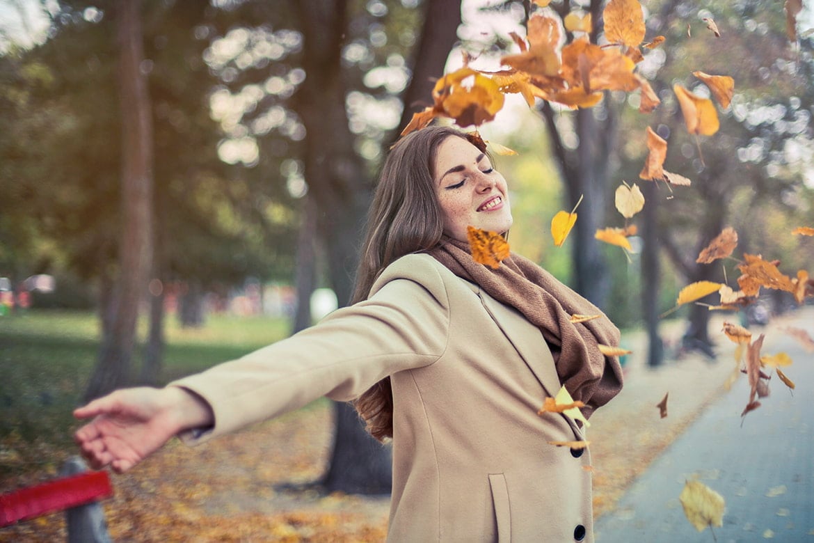 Junge Frau im Mantel wirft glücklich mit Herbstlaub