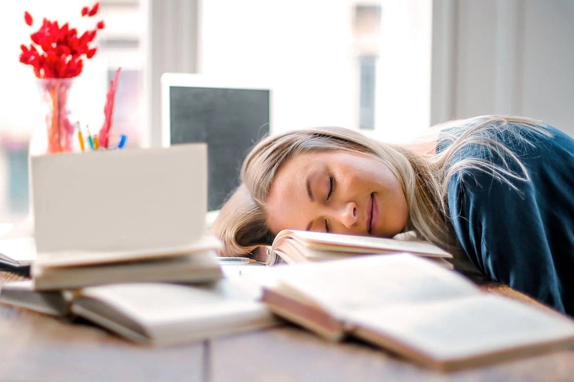Frau liegt schlafend mit dem Kopf auf Büchern am Arbeitstisch