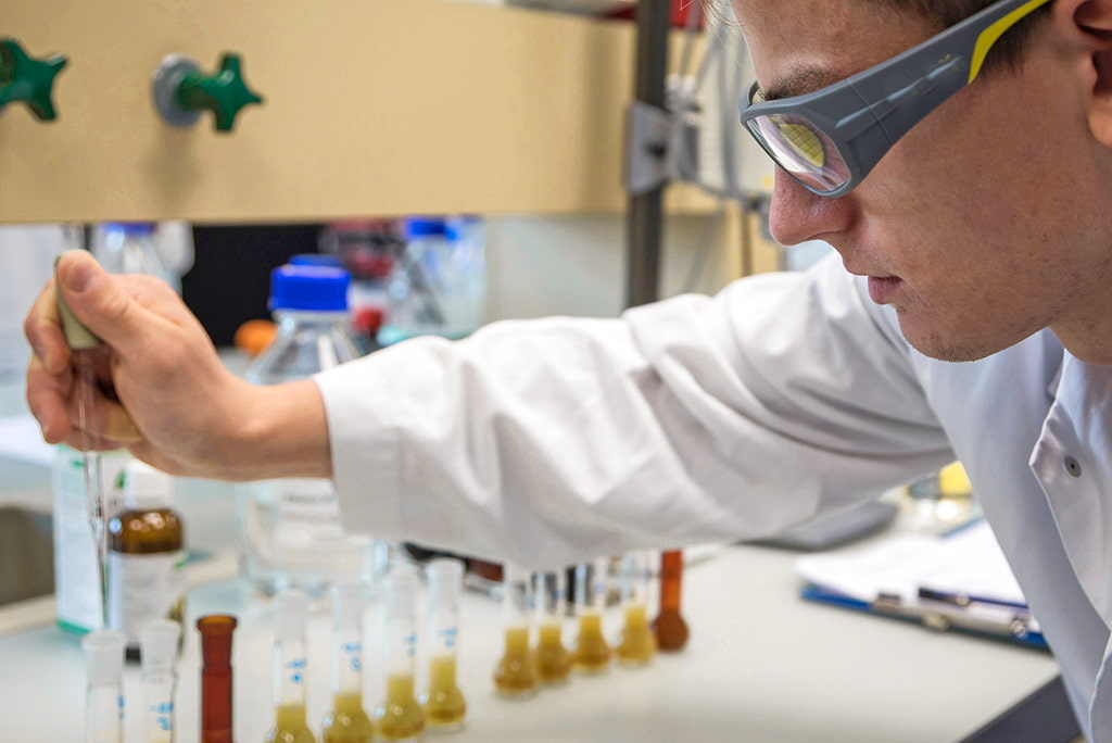 Junger Laborant in weissem Kittel und Schutzbrille arbeitet mit Pipette und pflanzlichen Substanzen