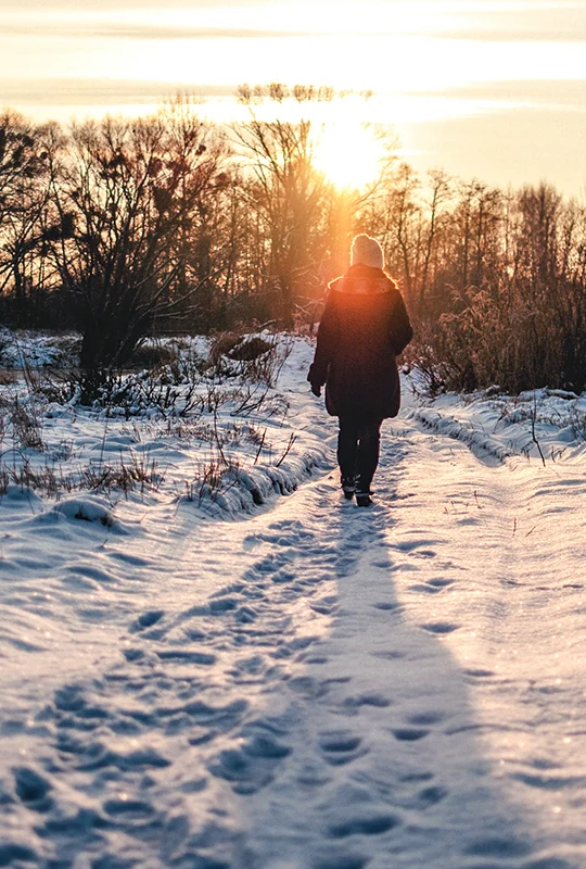 Eine Frau läuft in winterlicher Landschaft auf den abendlichen Sonnenuntergang zu