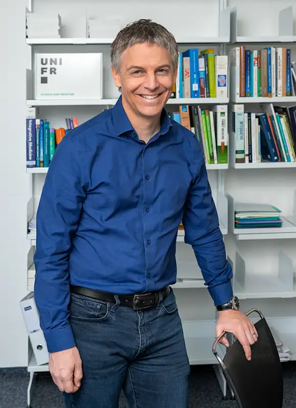 Portrait du Dr Pierre-Yves Rodondi, professeur ordinaire, Université de Fribourg, posant devant une bibliothèque, en chemise bleue.