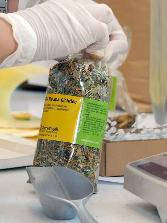 Hände in Gummihandschuhen füllen eine Tee-Hausmischung in eine Zellophan-Packung