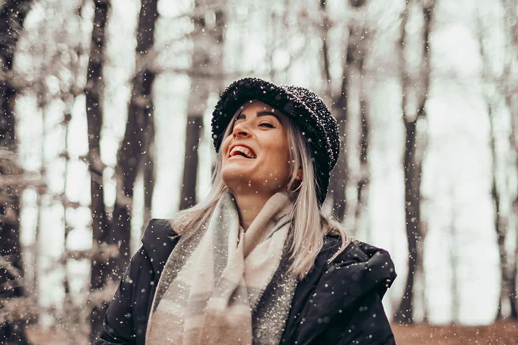 Lachende Frau steht mit Hut und Schal bekleidet im Schneetreiben im Wald